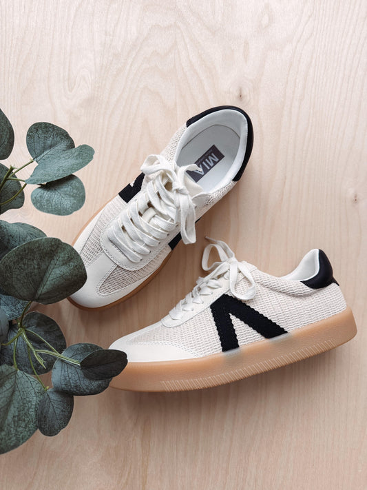 White sneakers with black accents on a light wooden floor with a plant.