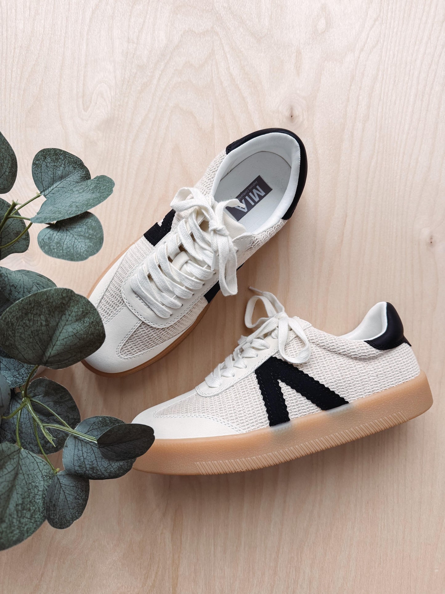 White sneakers with black accents on a light wooden floor with a plant.