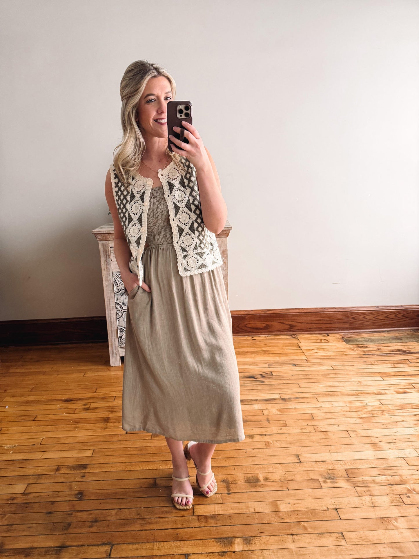 Woman taking a mirror selfie wearing a beige dress and white lace vest in a room with wooden flooring.