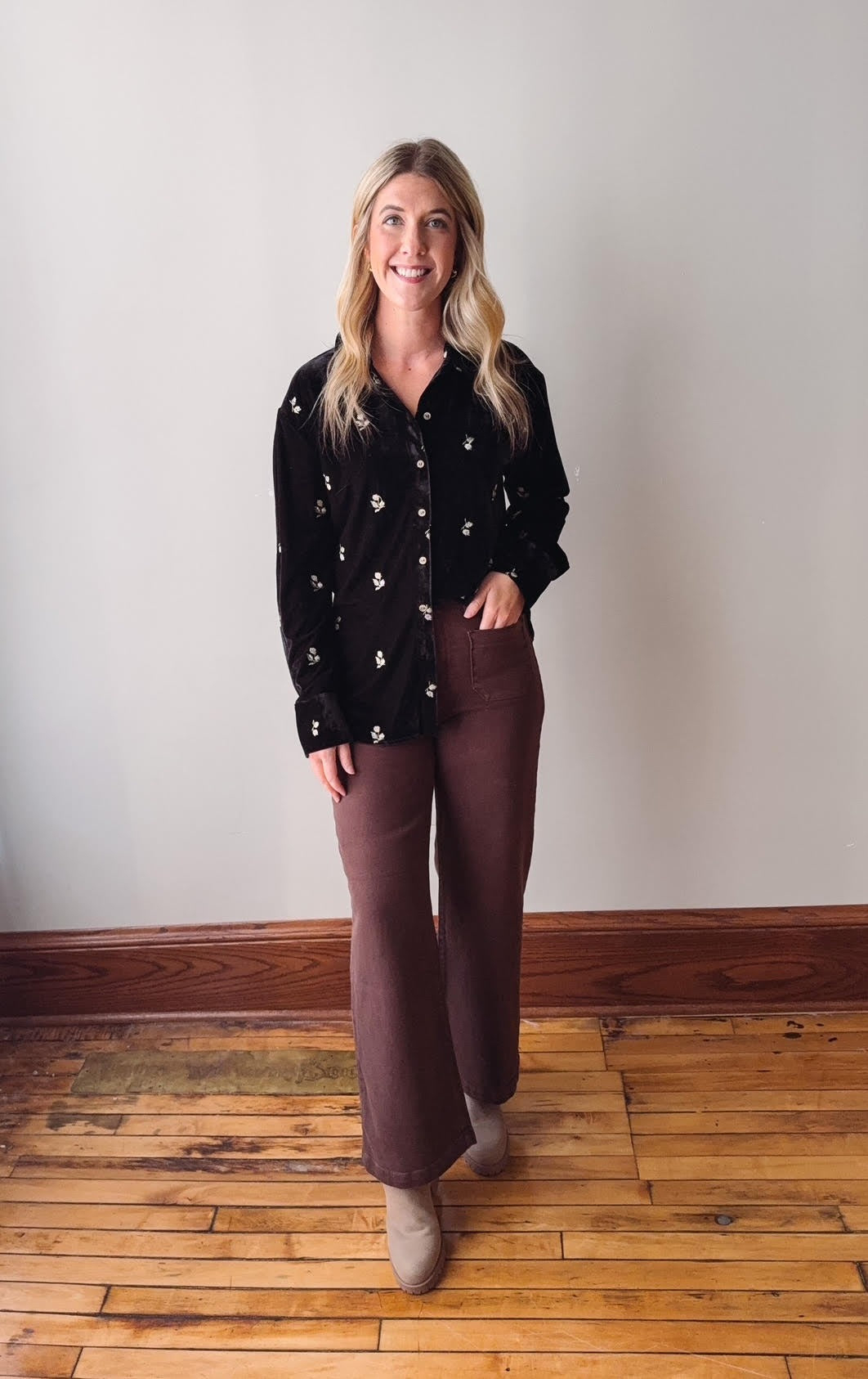 Woman wearing a black floral shirt and brown pants standing on a wooden floor.