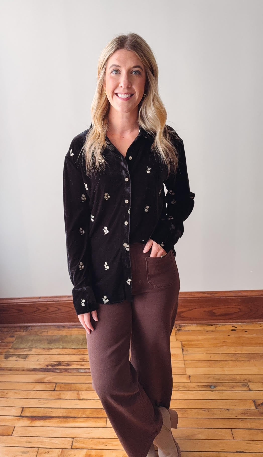 Woman wearing a black floral blouse and brown pants standing on a wooden floor.
