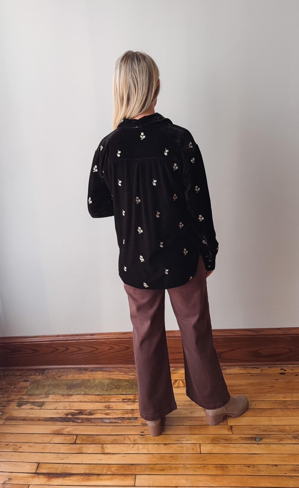 Person wearing a black patterned shirt and brown pants standing in a room with wooden flooring and white walls.