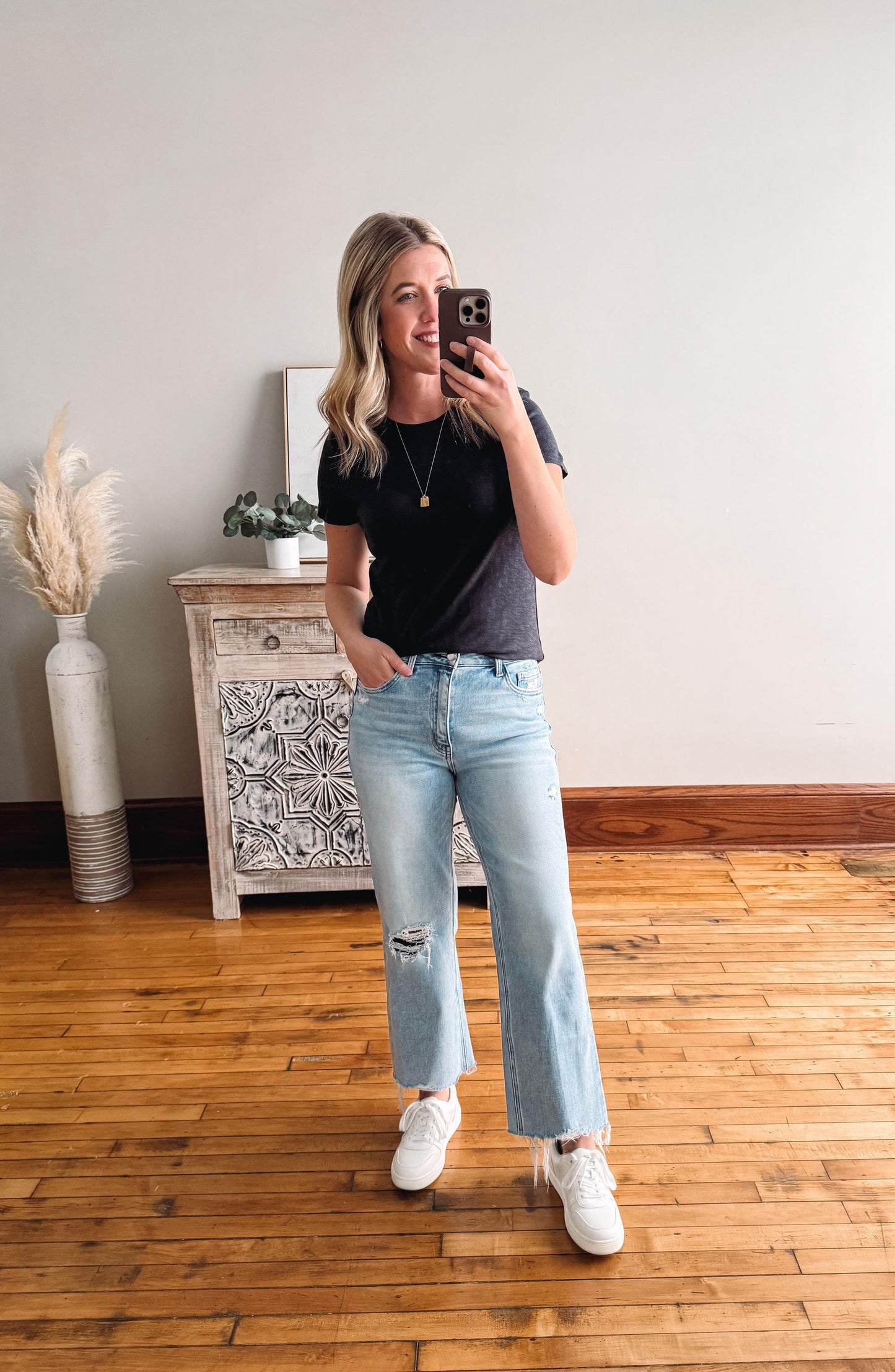 Person taking a mirror selfie wearing a black t-shirt, light blue jeans, and white sneakers in a room with wooden flooring and a decorative cabinet.