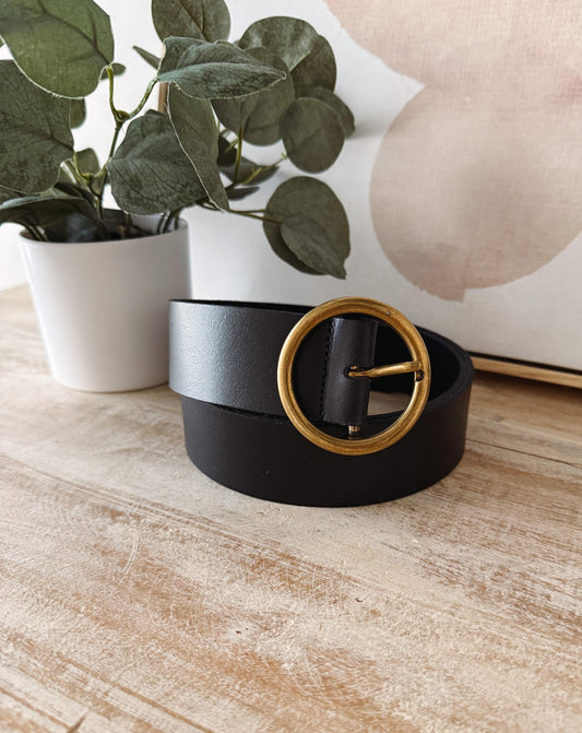 Black belt with a gold buckle on a wooden surface with a plant in the background