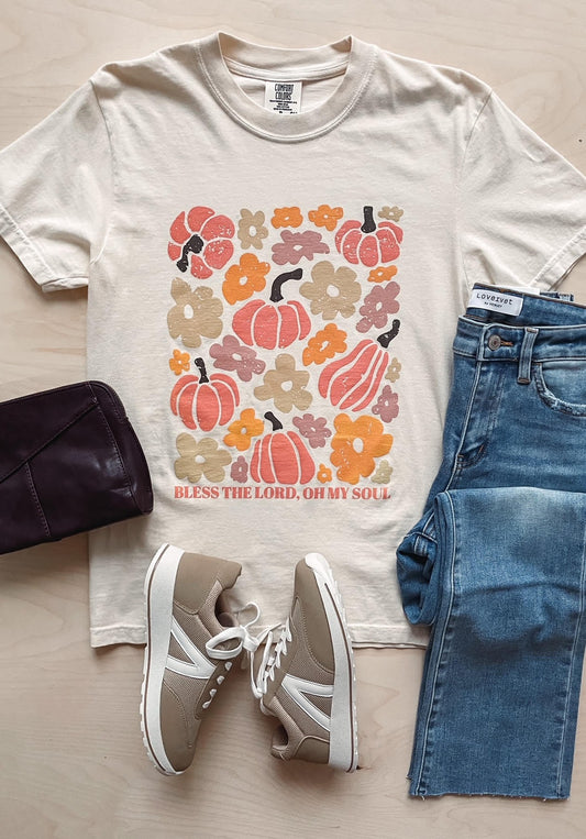 A flat lay of a beige Comfort Colors unisex t-shirt with a pumpkin and abstract floral graphic, the text 'Bless the Lord, Oh my soul,' and accompanied by a pair of blue jeans and a pair of beige sneakers.