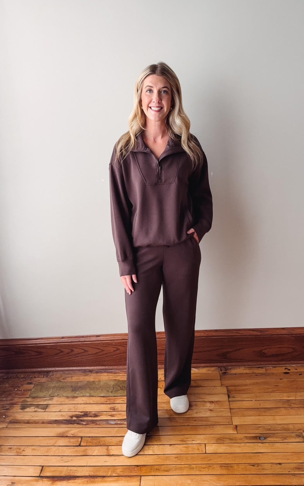 Woman wearing a brown outfit standing on a wooden floor against a white wall