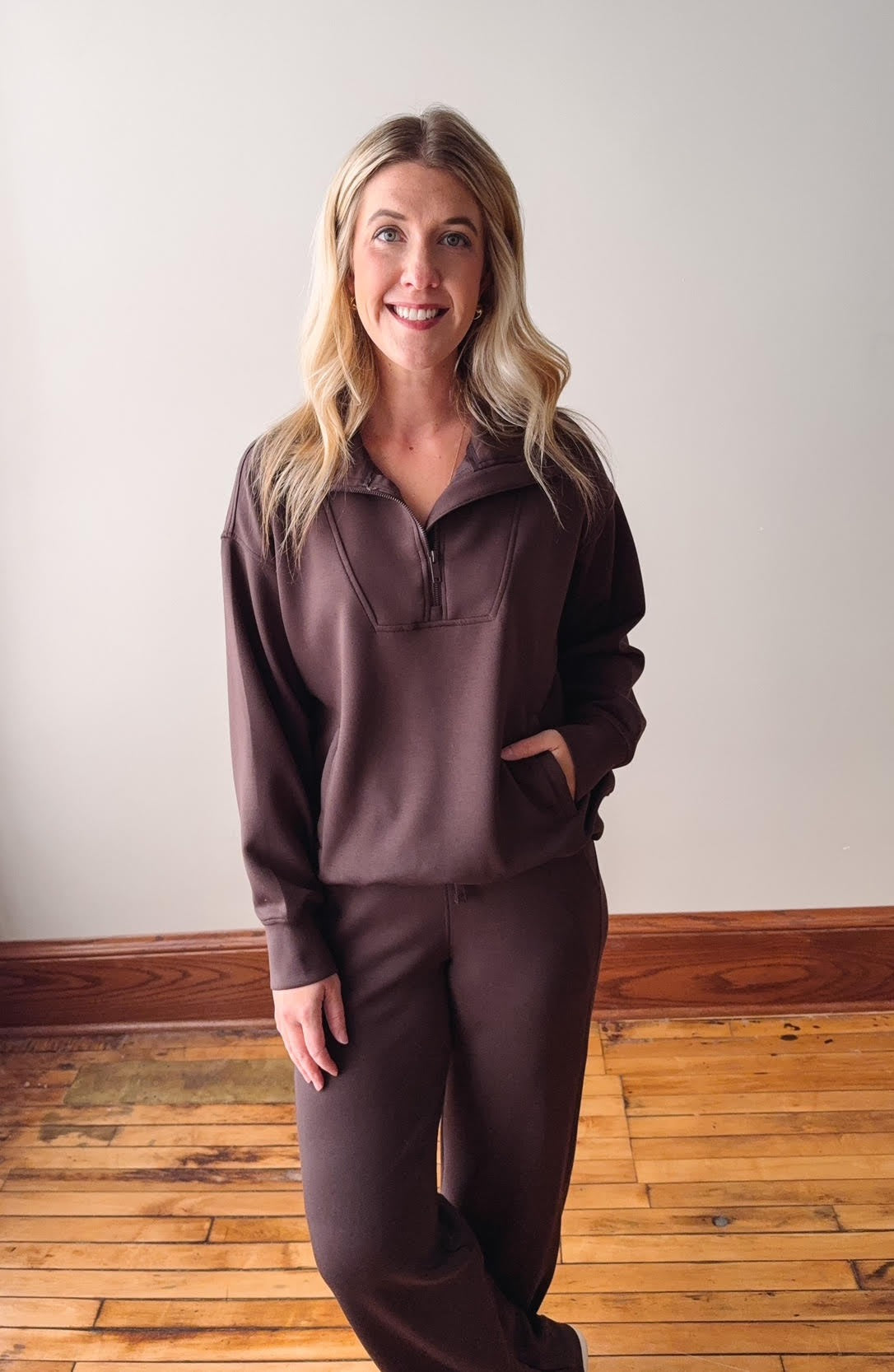 Woman wearing a brown outfit standing on a wooden floor with a white wall background
