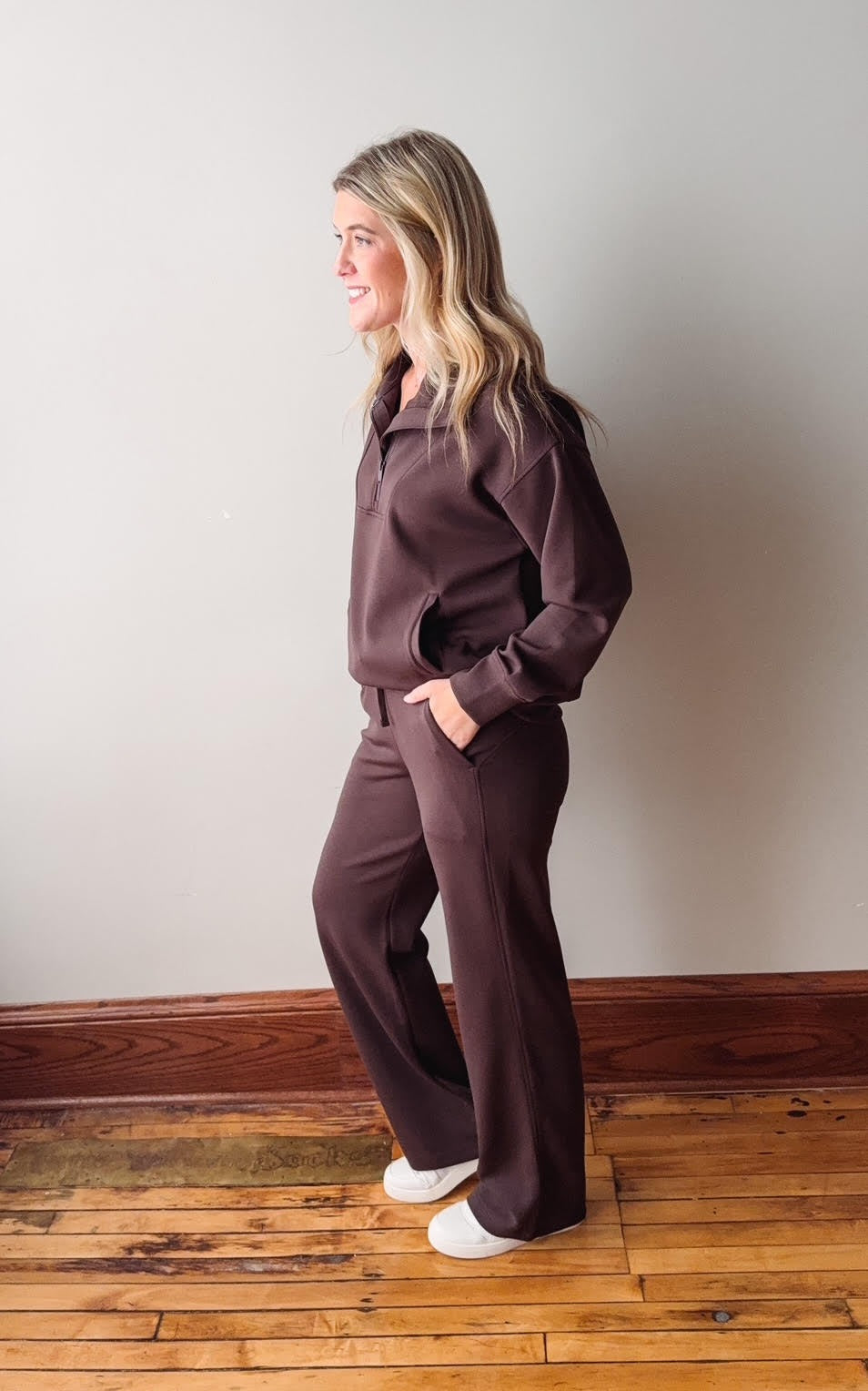 Woman wearing a brown outfit standing against a plain wall on a wooden floor.