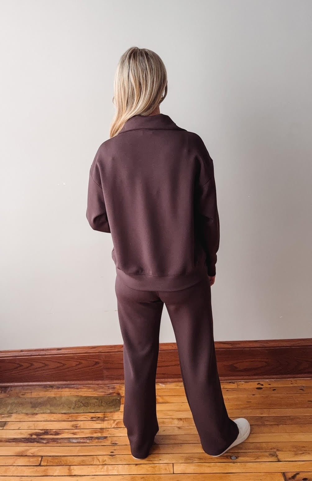 Person wearing a brown tracksuit standing on a wooden floor with a white wall background