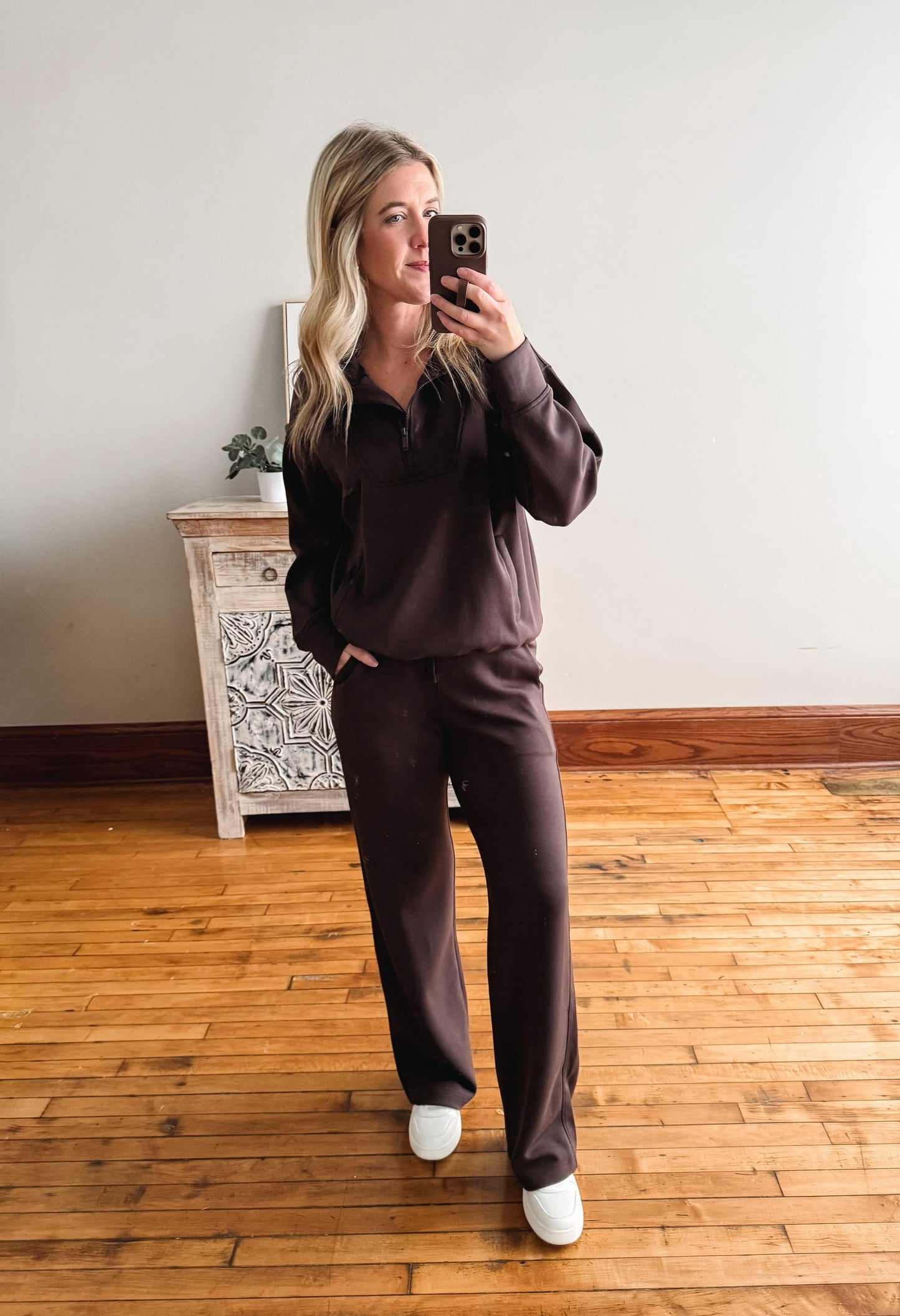 Person wearing a brown outfit taking a mirror selfie in a room with wooden flooring and a decorative cabinet.