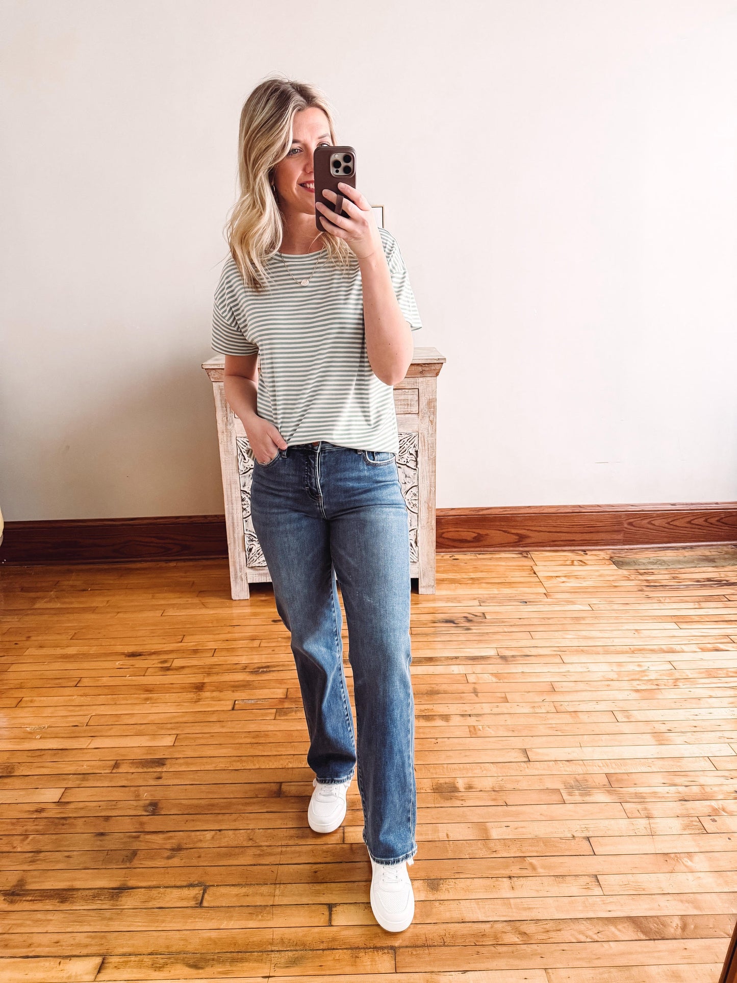 Woman taking a mirror selfie wearing a striped shirt and jeans in a room with wooden flooring.