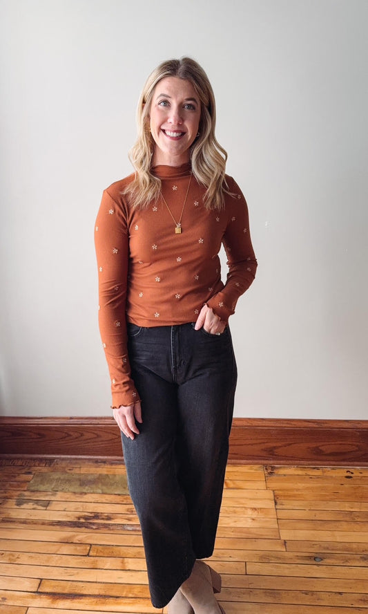 Woman wearing a rust-colored long-sleeve top with black pants on a wooden floor.