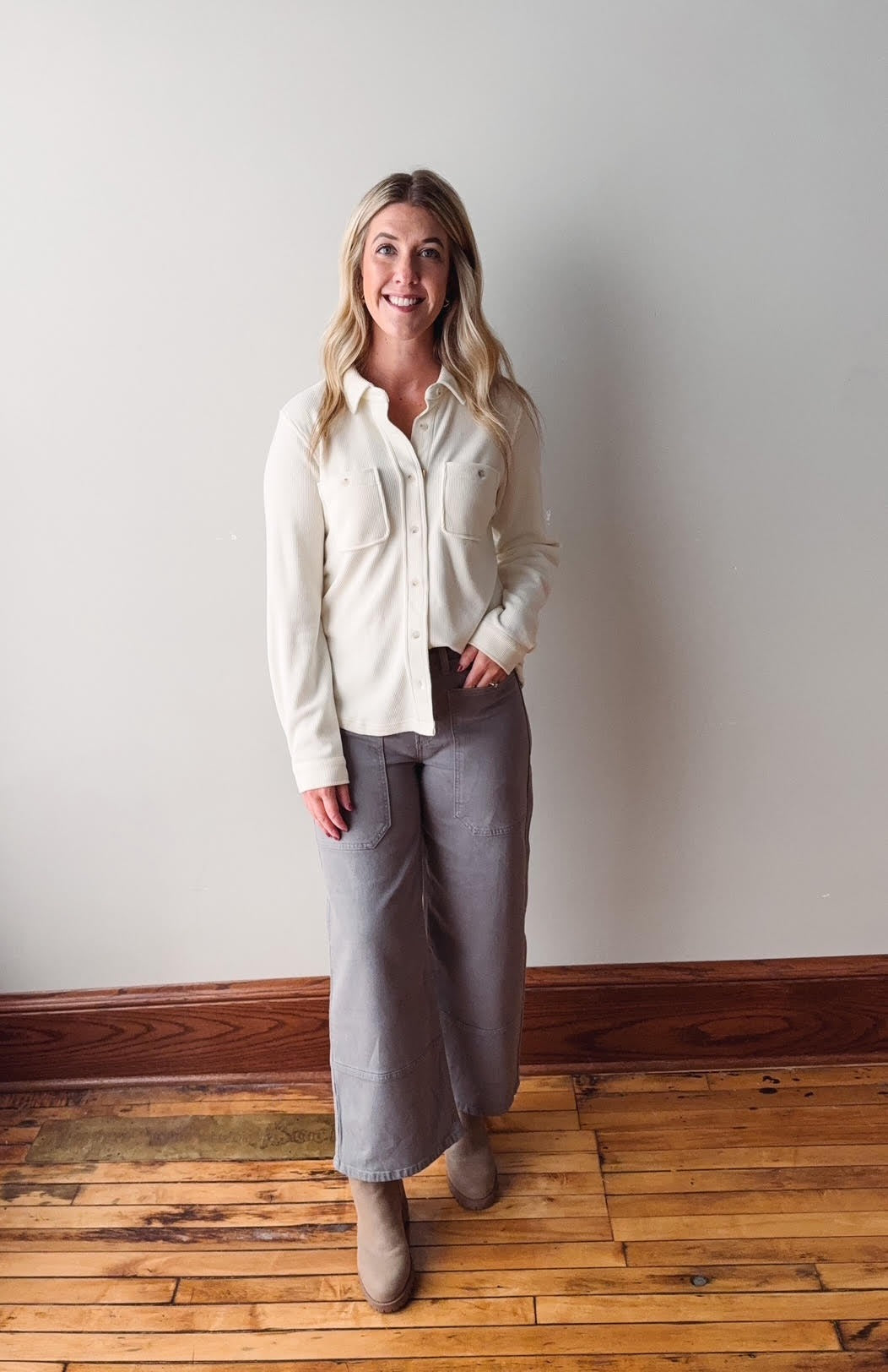 Woman wearing a white blouse and gray pants standing against a plain wall.