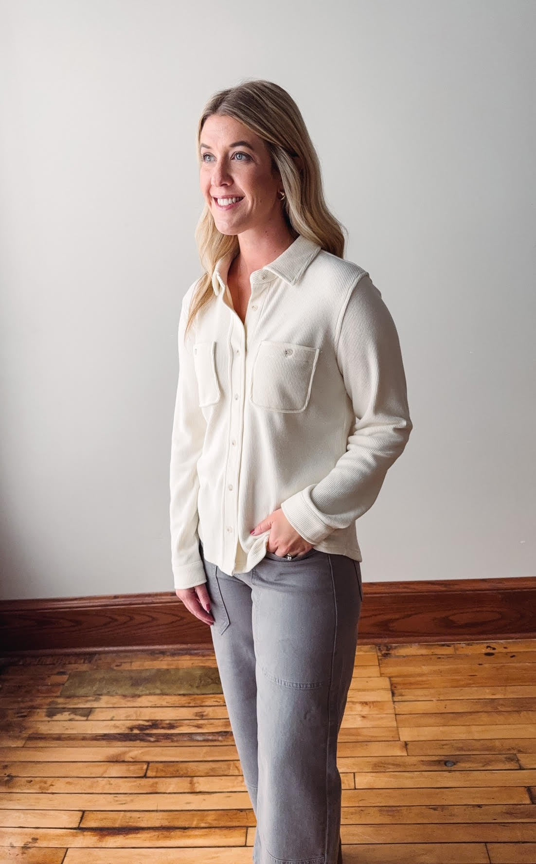 Woman wearing a white blouse and gray pants standing on a wooden floor with a white wall background