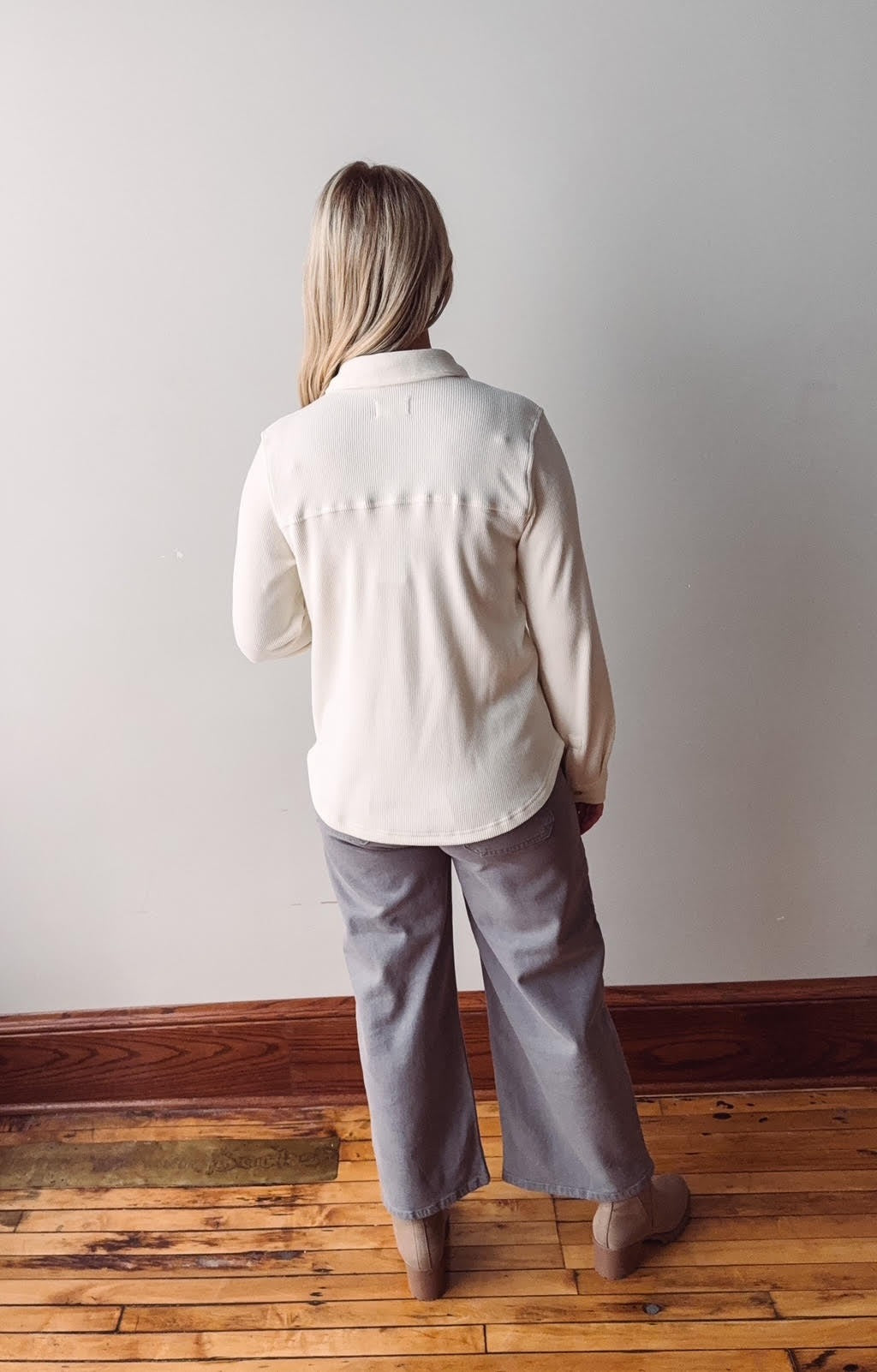 Person wearing a white shirt and gray pants standing in a room with wooden flooring and a white wall.