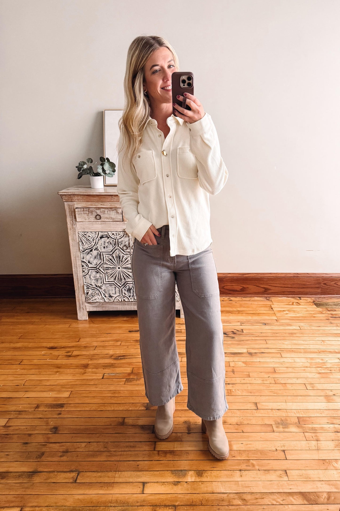 Woman taking a mirror selfie wearing a white shirt and gray pants in a room with wooden flooring and a decorative cabinet.