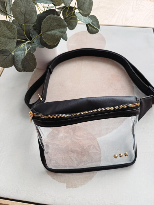 Clear fanny pack with black strap and gold zipper on a light surface with greenery.