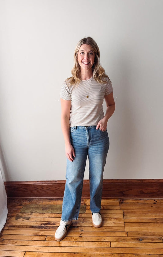 Woman wearing a beige t-shirt and blue jeans standing on a wooden floor against a white wall.