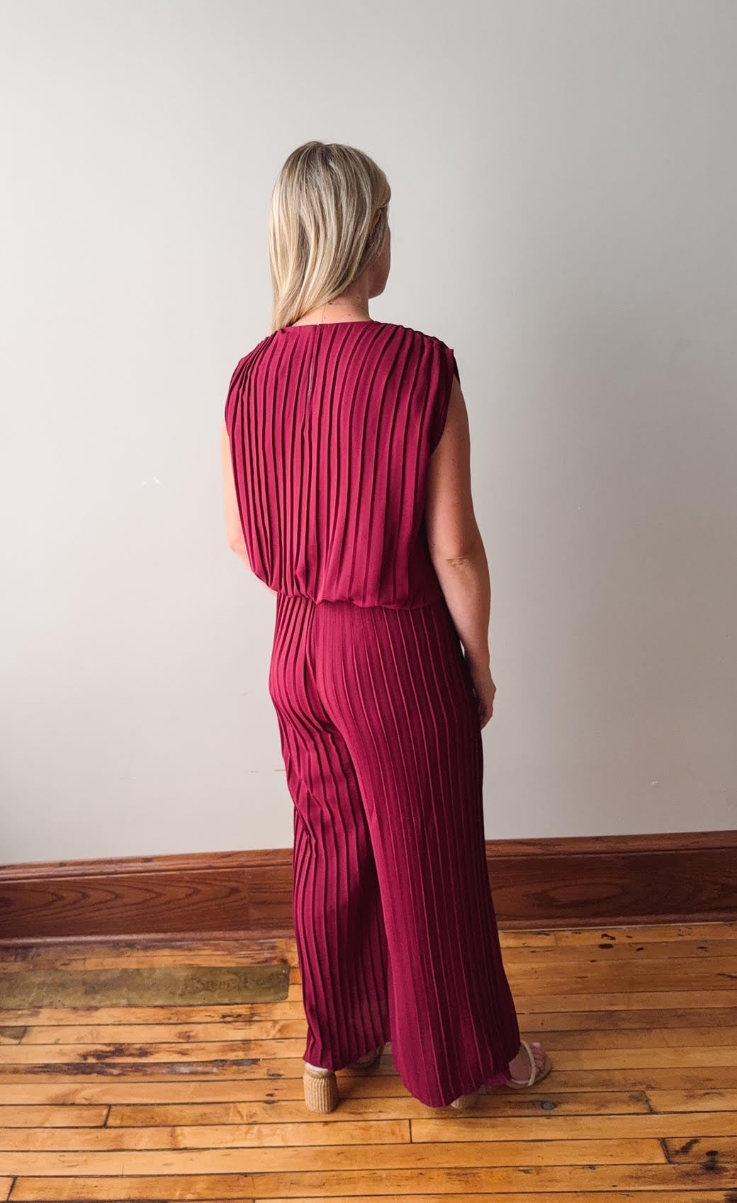 Woman wearing a maroon pleated jumpsuit standing on a wooden floor with a white wall background