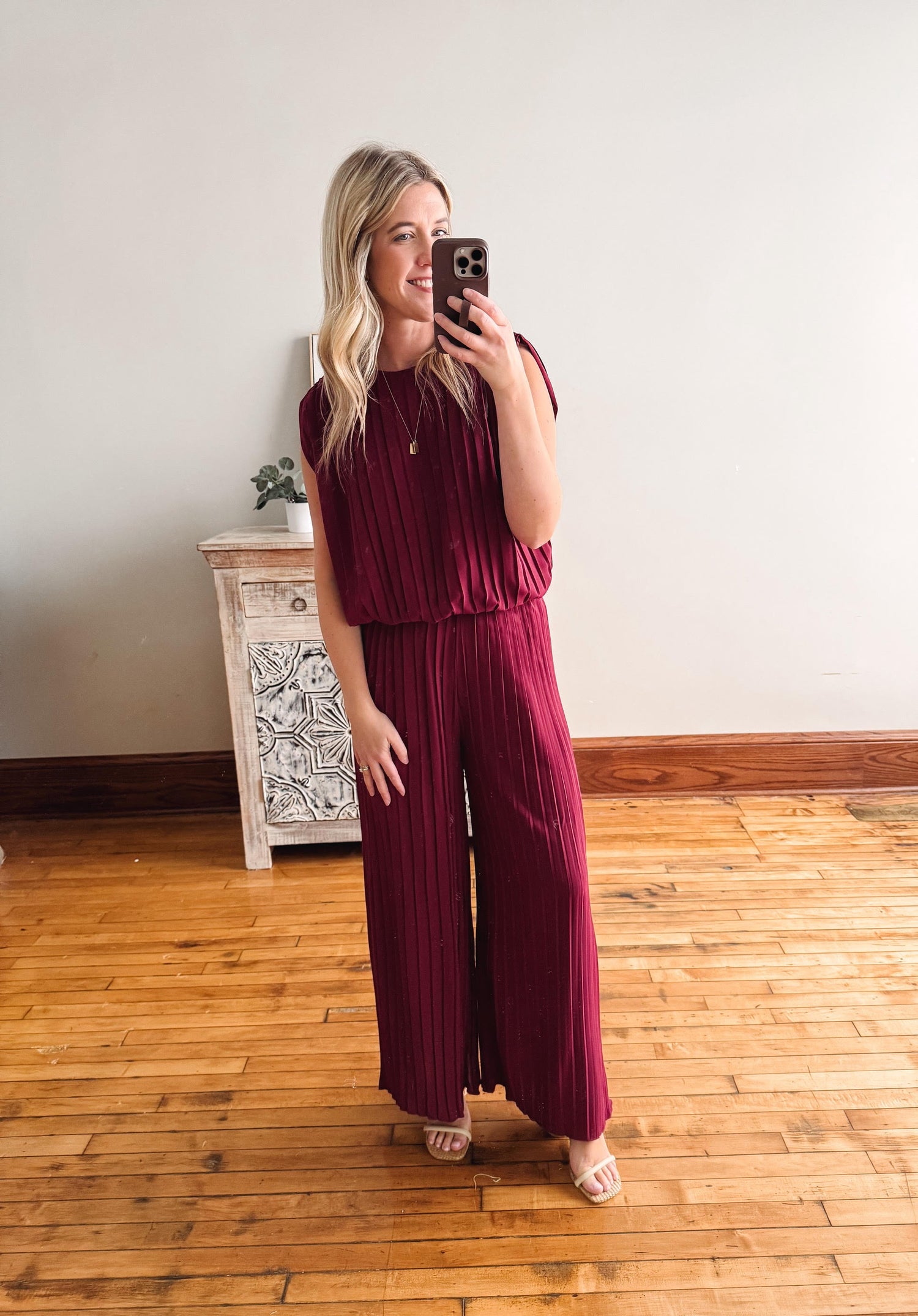 Woman taking a mirror selfie wearing a burgundy jumpsuit in a room with wooden flooring and a decorative cabinet.