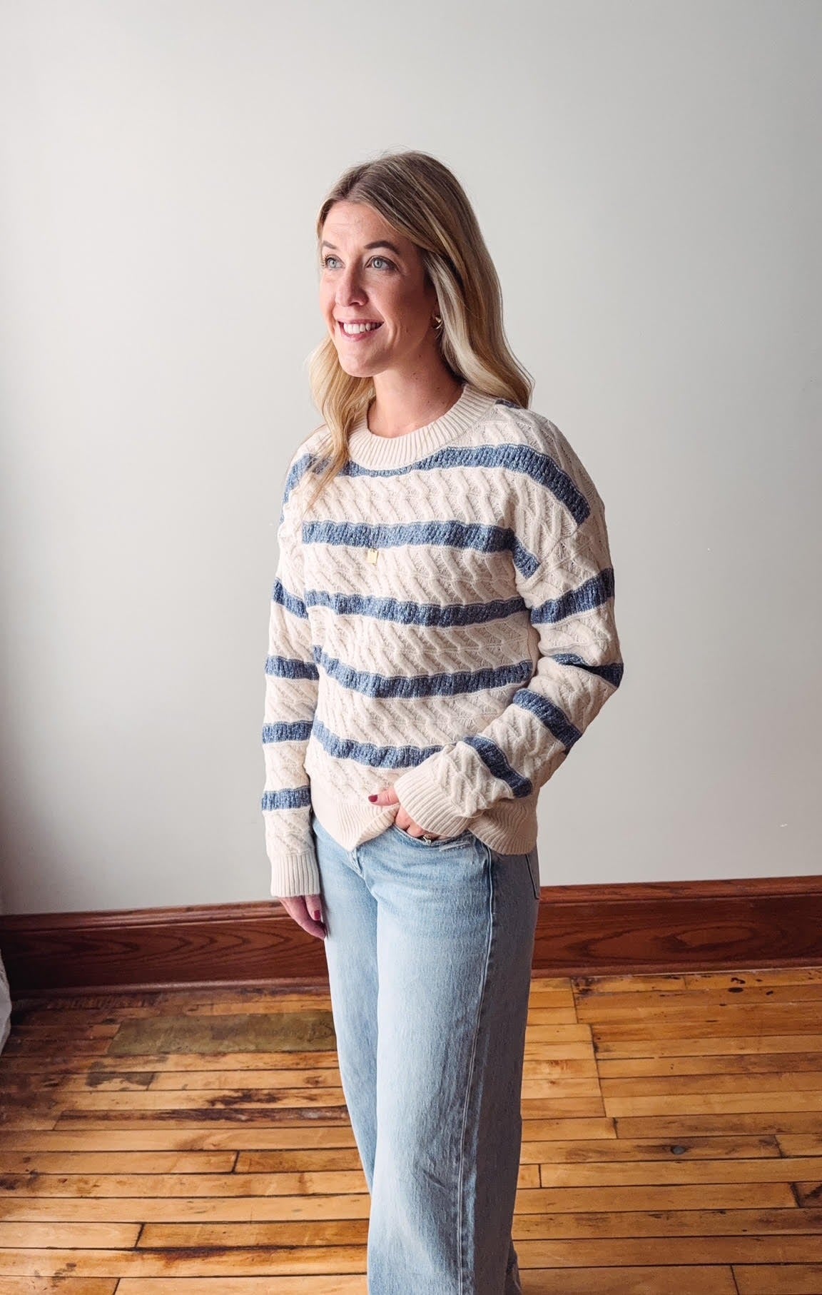 Woman wearing a striped sweater and jeans standing in a room with wooden flooring and a white wall.