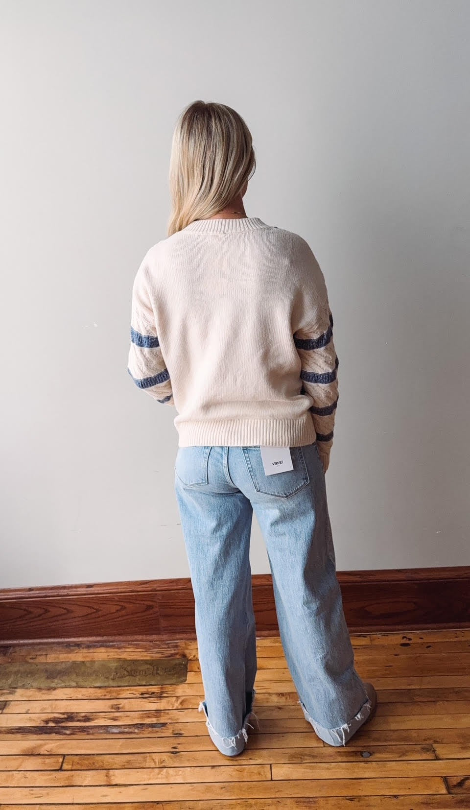 Person wearing a light-colored sweater with dark stripes and light blue jeans, standing on a wooden floor against a plain wall.
