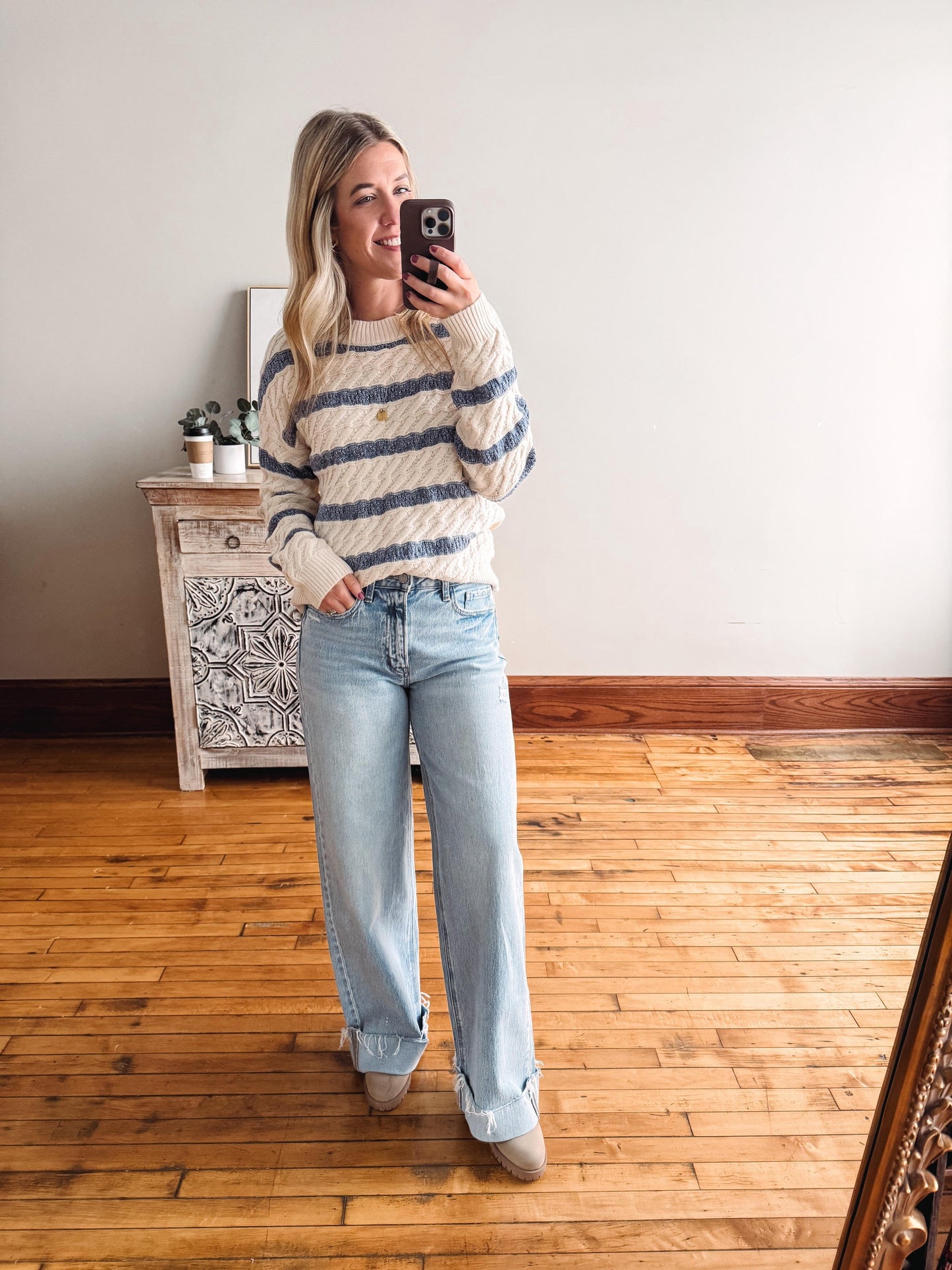 Woman taking a mirror selfie wearing a striped sweater and jeans in a room with wooden flooring.