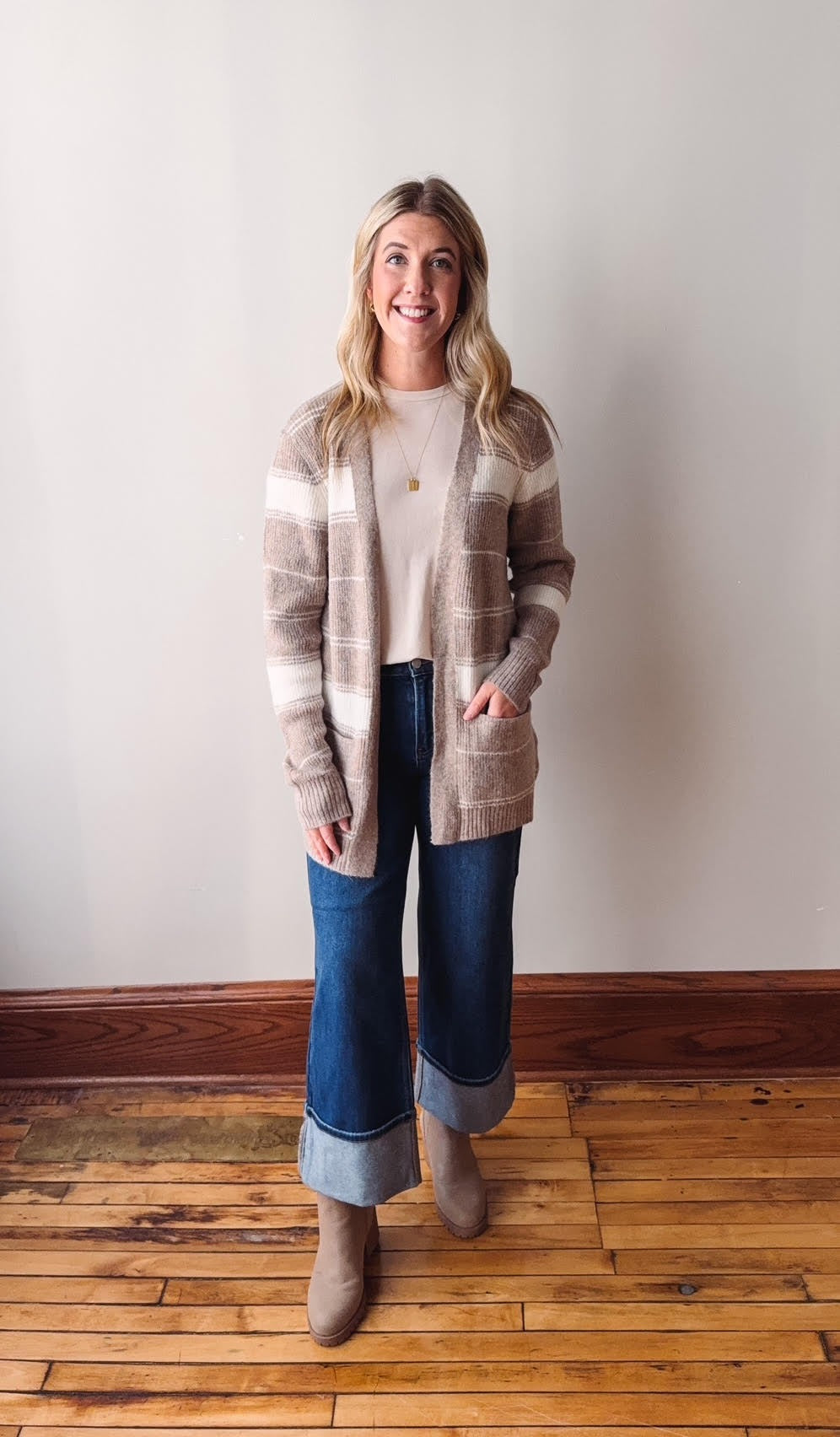 Woman wearing a striped cardigan and jeans standing on a wooden floor with a white wall background