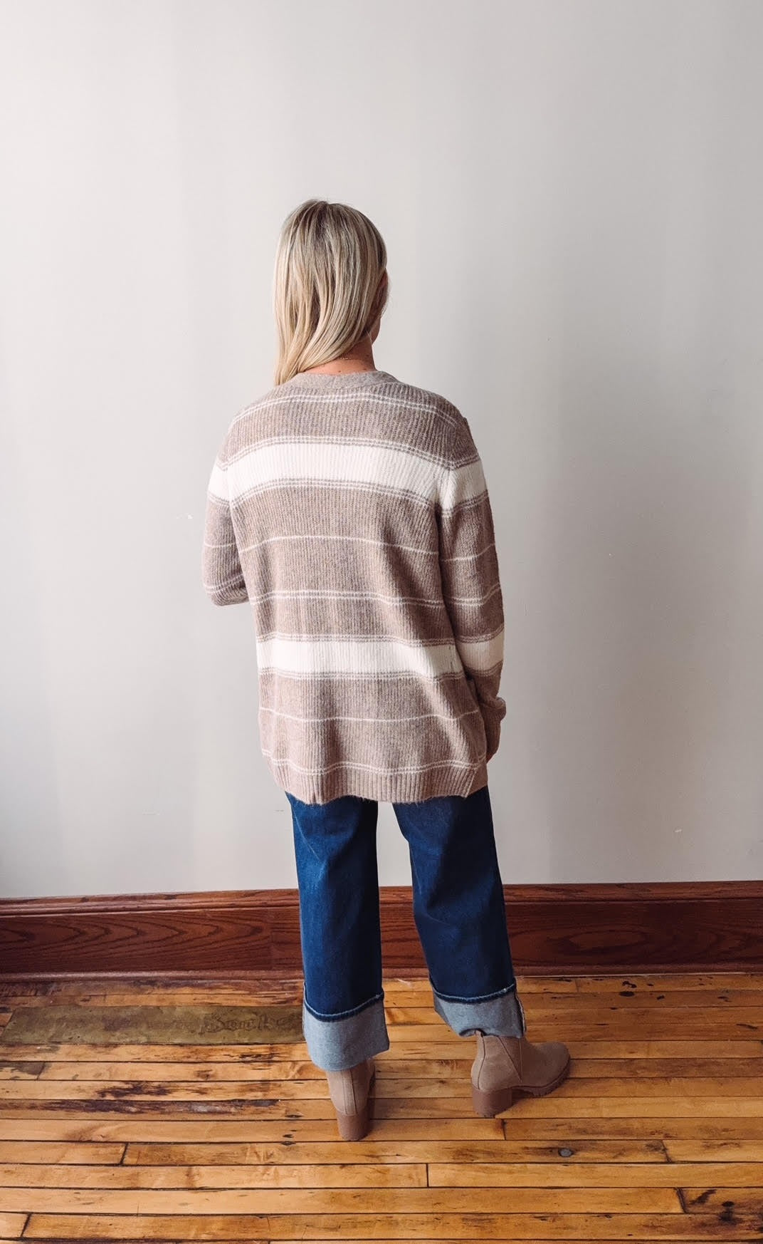 Person wearing a striped sweater and jeans standing on a wooden floor against a white wall.