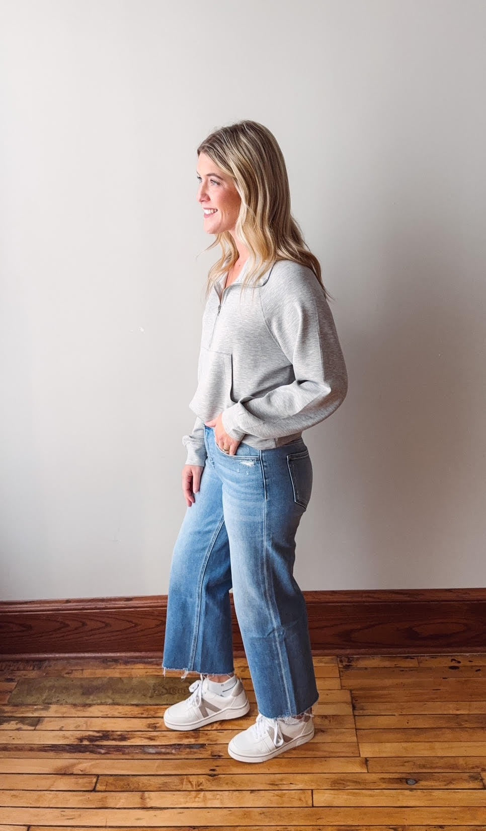 Woman wearing a gray sweater and blue jeans standing on a wooden floor against a white wall.