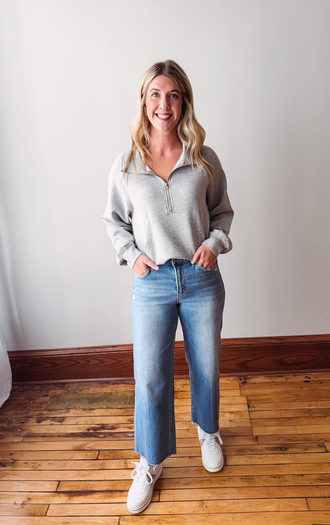 Woman wearing a gray sweater, blue jeans, and white sneakers standing on a wooden floor.