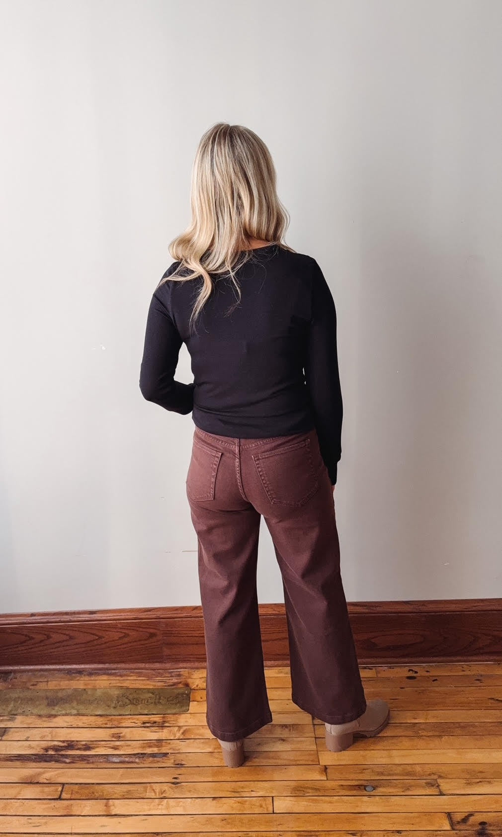 Person wearing a black top and brown pants standing against a plain wall.