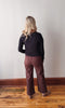 Person wearing a black top and brown pants standing against a plain wall.