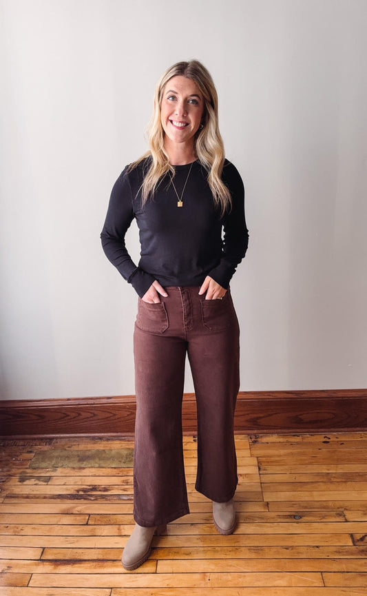 Woman wearing a black top and brown pants standing on a wooden floor with a white wall background