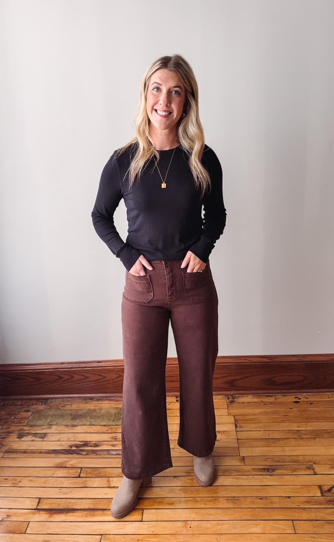Woman wearing a black top and brown pants standing on a wooden floor with a white wall background
