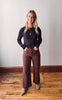 Woman wearing a black top and brown pants standing on a wooden floor with a white wall background
