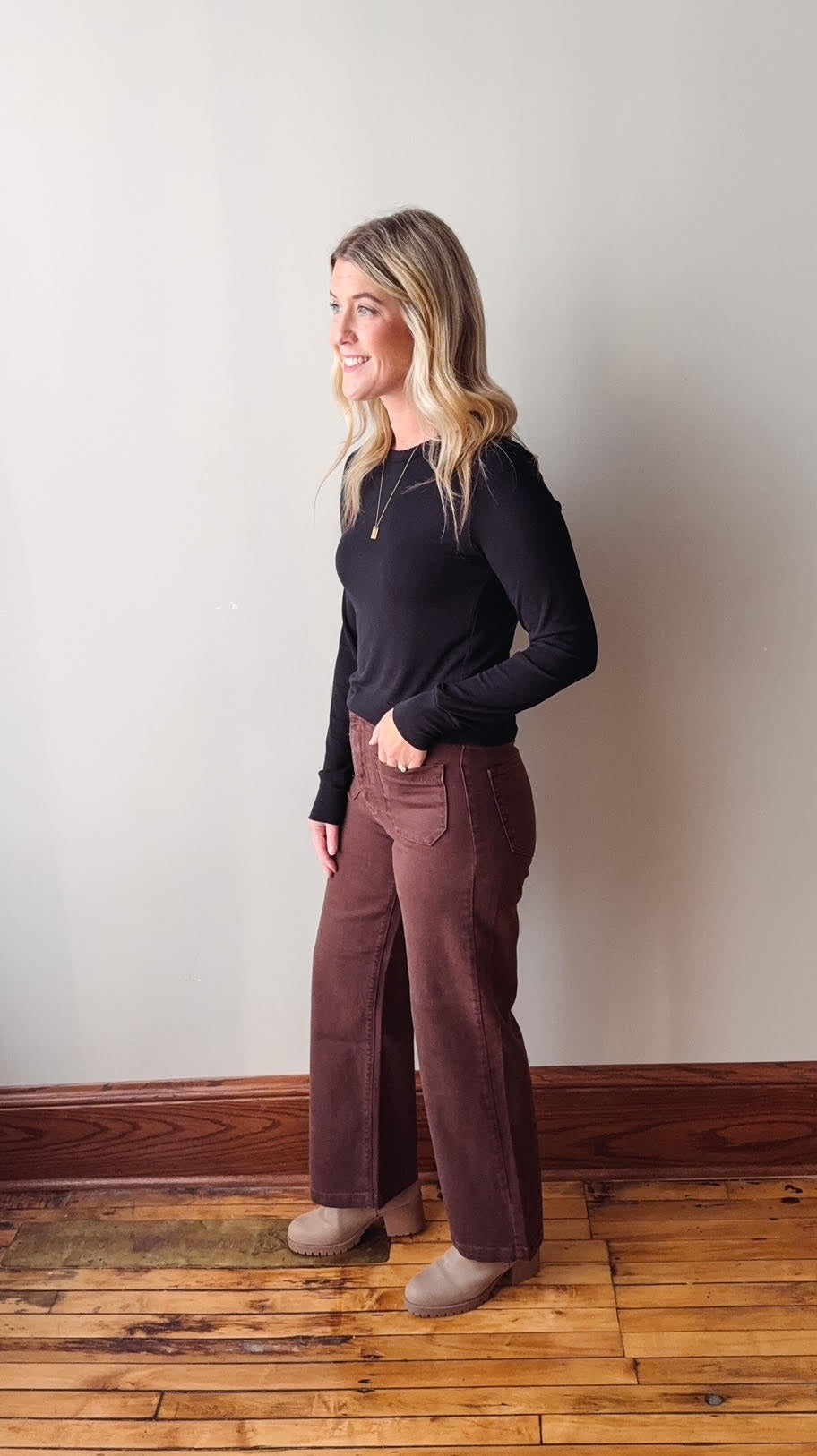 Woman wearing a black top and brown pants standing against a white wall.