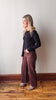 Woman wearing a black top and brown pants standing against a white wall.
