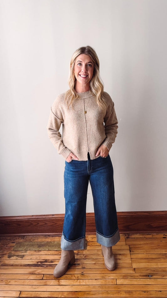 Woman wearing a beige sweater and blue jeans standing against a plain wall.