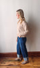 Woman wearing a beige sweater and blue jeans standing against a white wall.