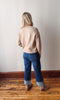 Person wearing a beige sweater and blue jeans standing against a plain wall.