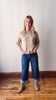 Woman wearing a beige sweater and blue jeans standing against a plain wall.