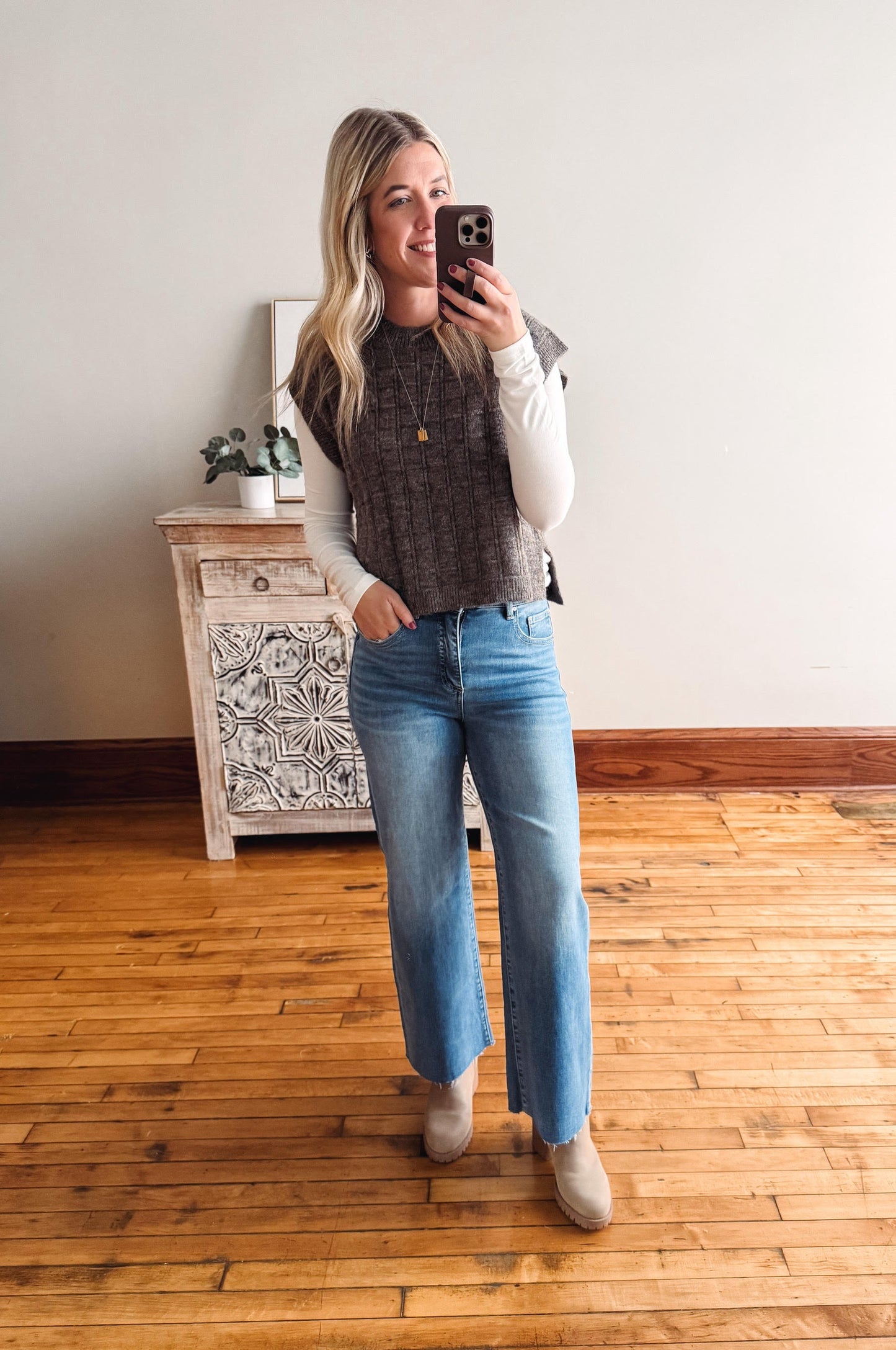 Woman taking a mirror selfie wearing a sweater, vest, and jeans in a room with wooden flooring and a decorative cabinet.