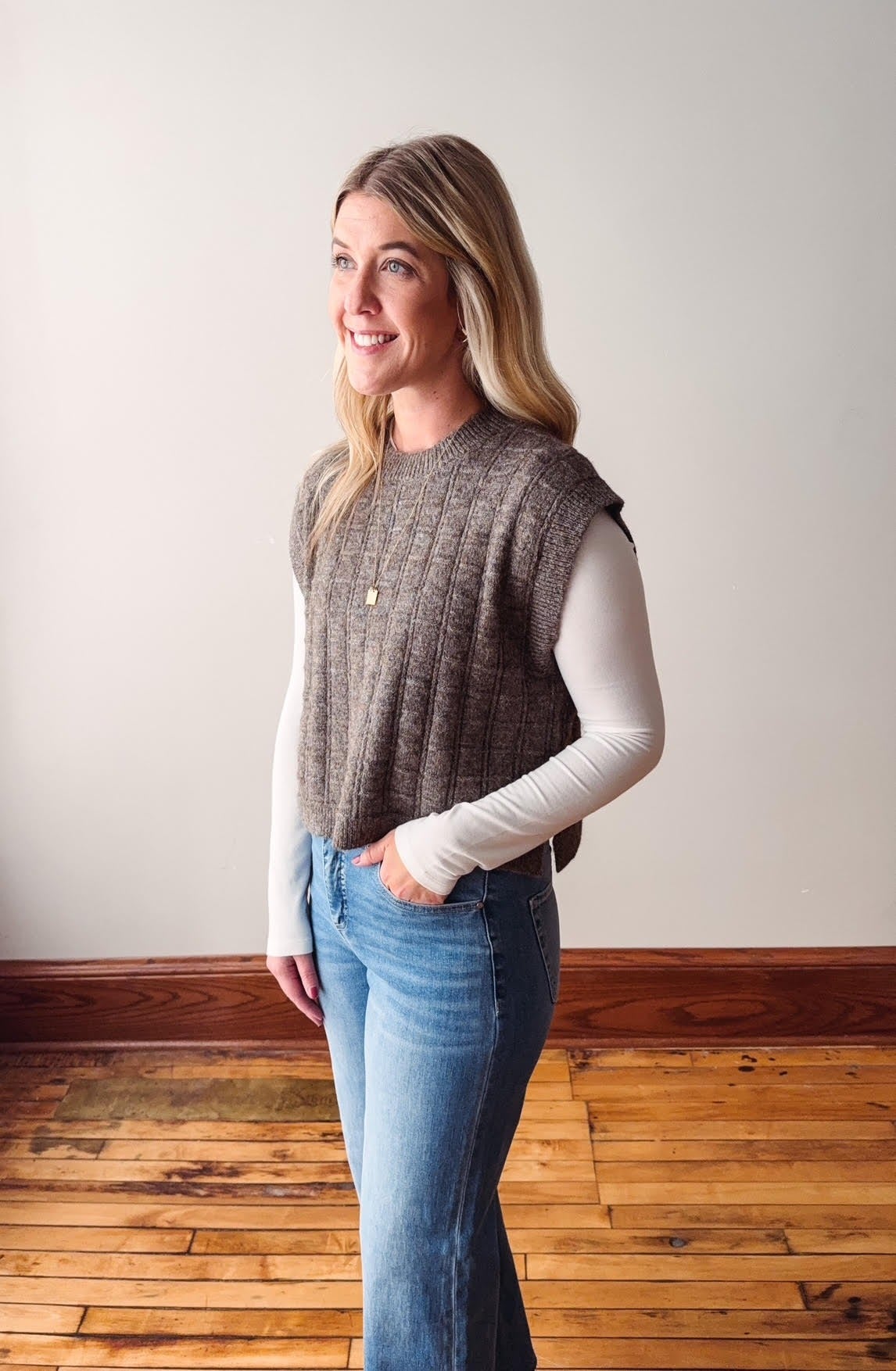 Woman wearing a brown knitted vest over a white long-sleeve shirt and blue jeans, standing against a plain wall.