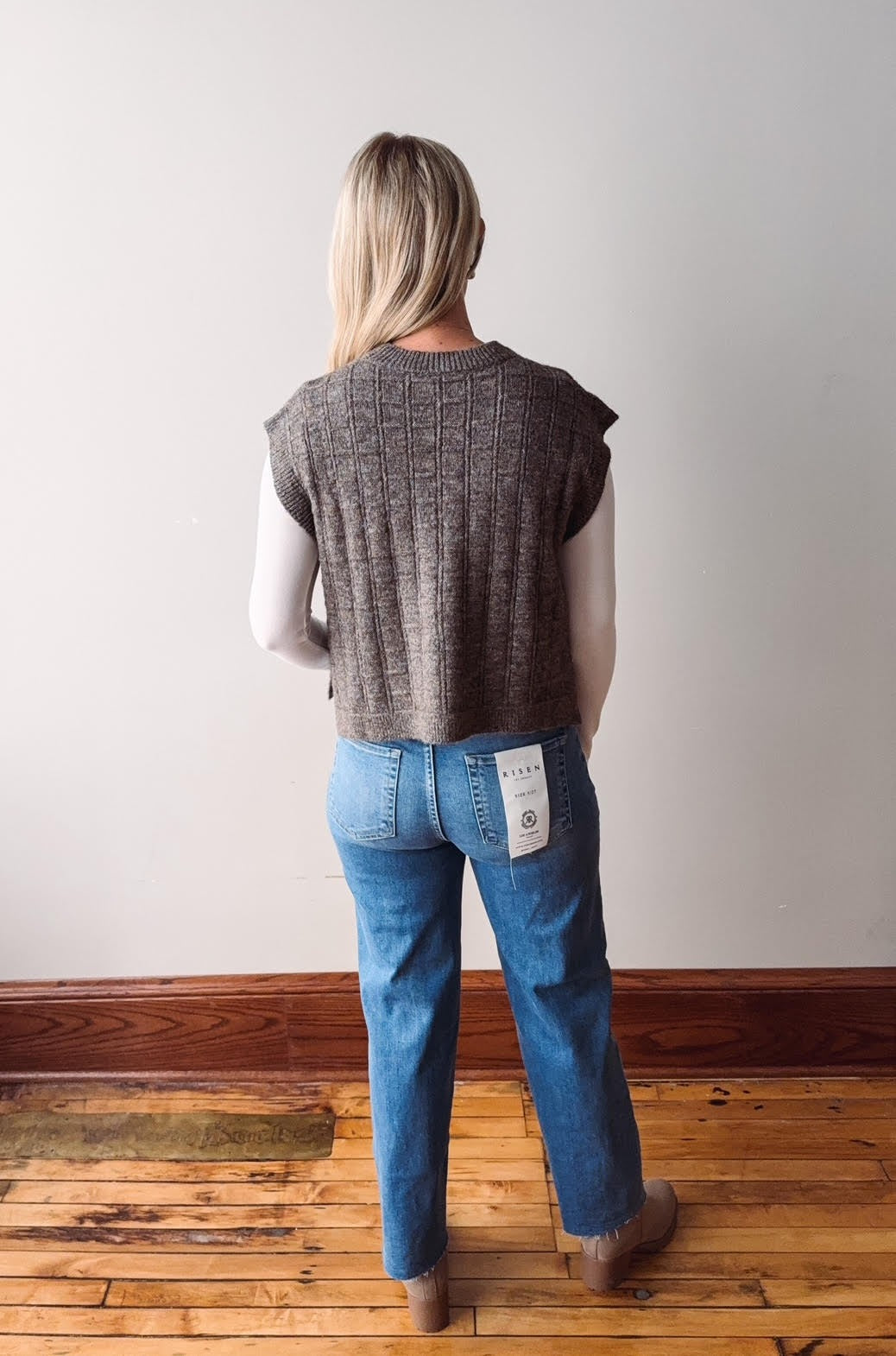Person wearing a sleeveless brown top and blue jeans standing on a wooden floor with a white wall background.