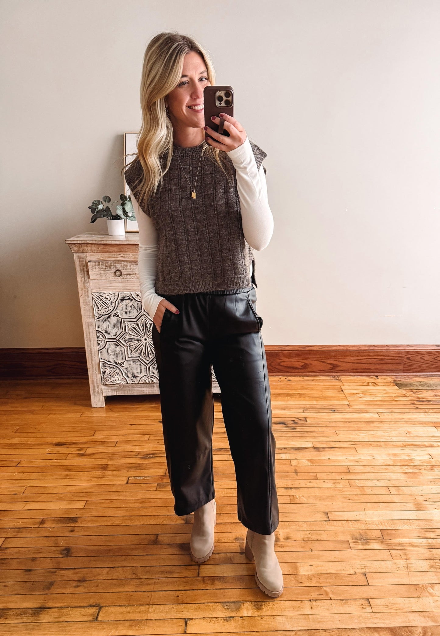 Woman taking a mirror selfie wearing a brown sweater vest, white shirt, black pants, and beige boots in a room with wooden flooring.