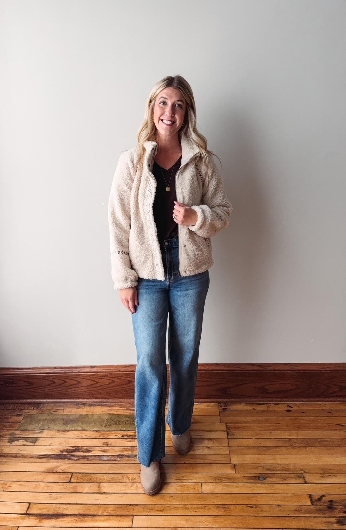Woman wearing a beige fleece jacket and blue jeans standing against a plain wall.
