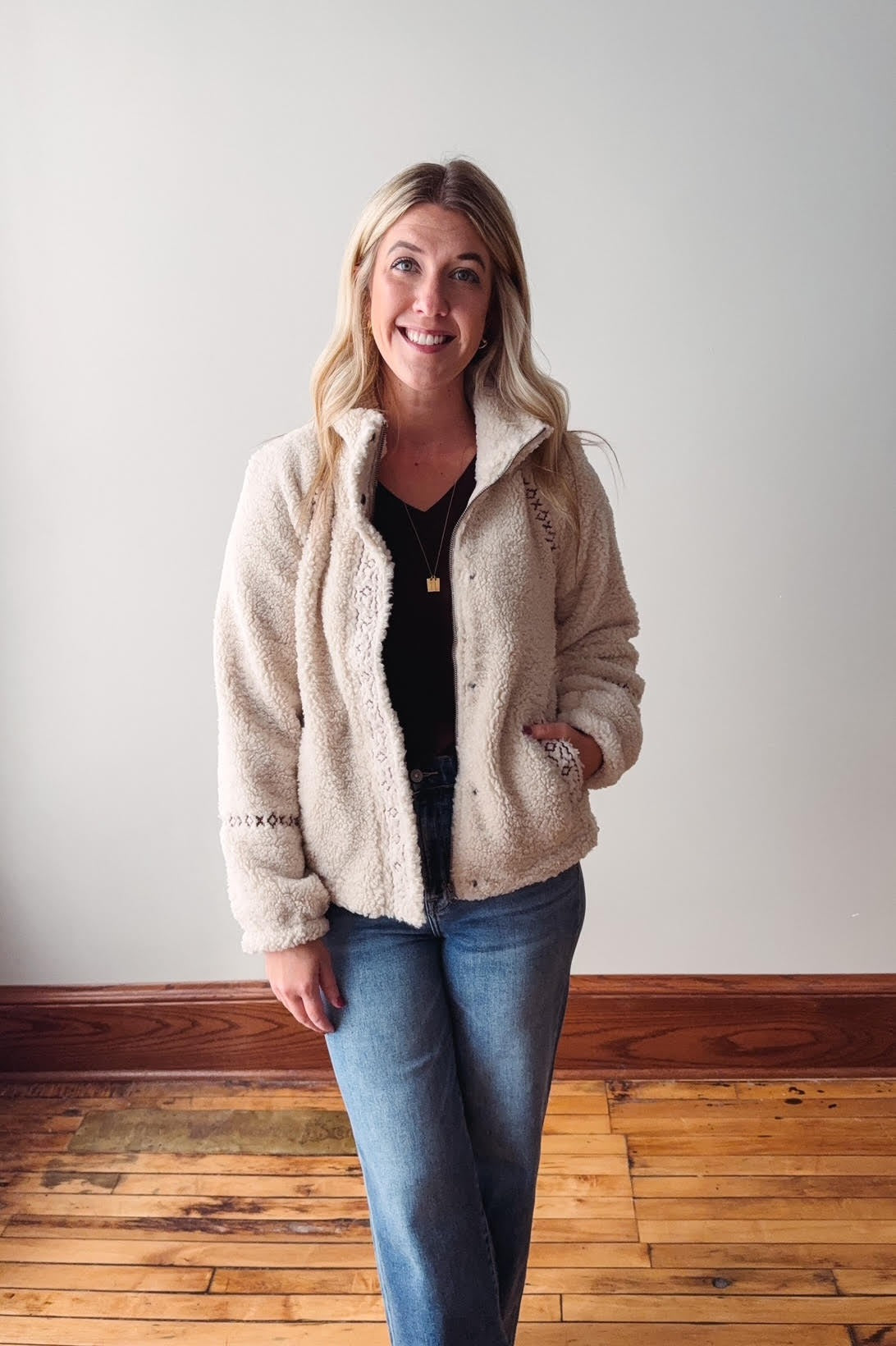 Woman wearing a beige sherpa jacket and blue jeans standing against a plain wall.
