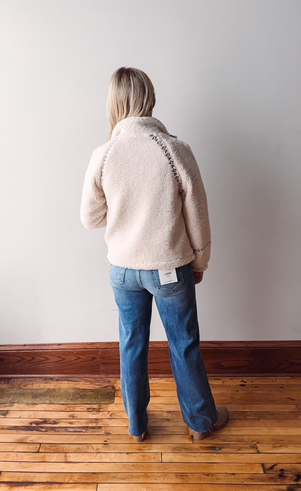 Person wearing a light pink jacket and blue jeans standing against a plain wall.
