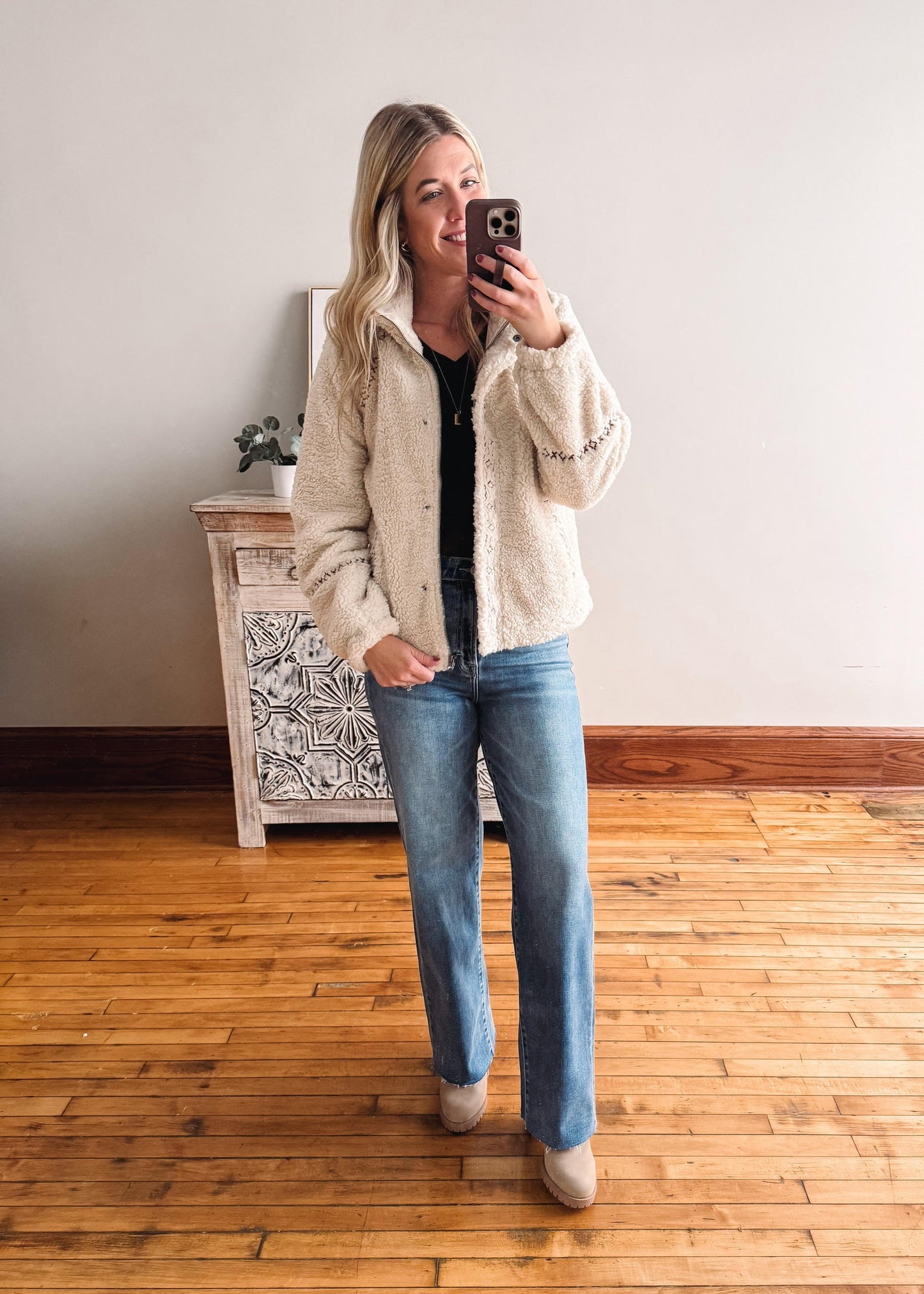 Woman taking a mirror selfie wearing a beige jacket and blue jeans in a room with wooden flooring.